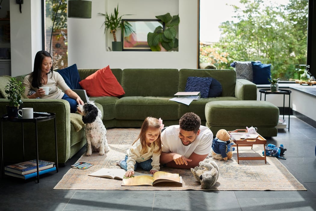 A parent and a small child reading together on a living room rug while another parent works from the sofa.